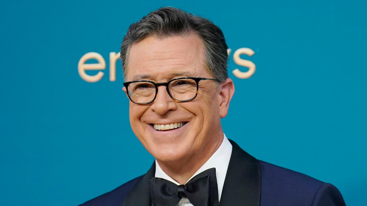 Moderator Stephen Colbert steht vor einer blauen Wand, er trägt eine Brille und lächelt.
