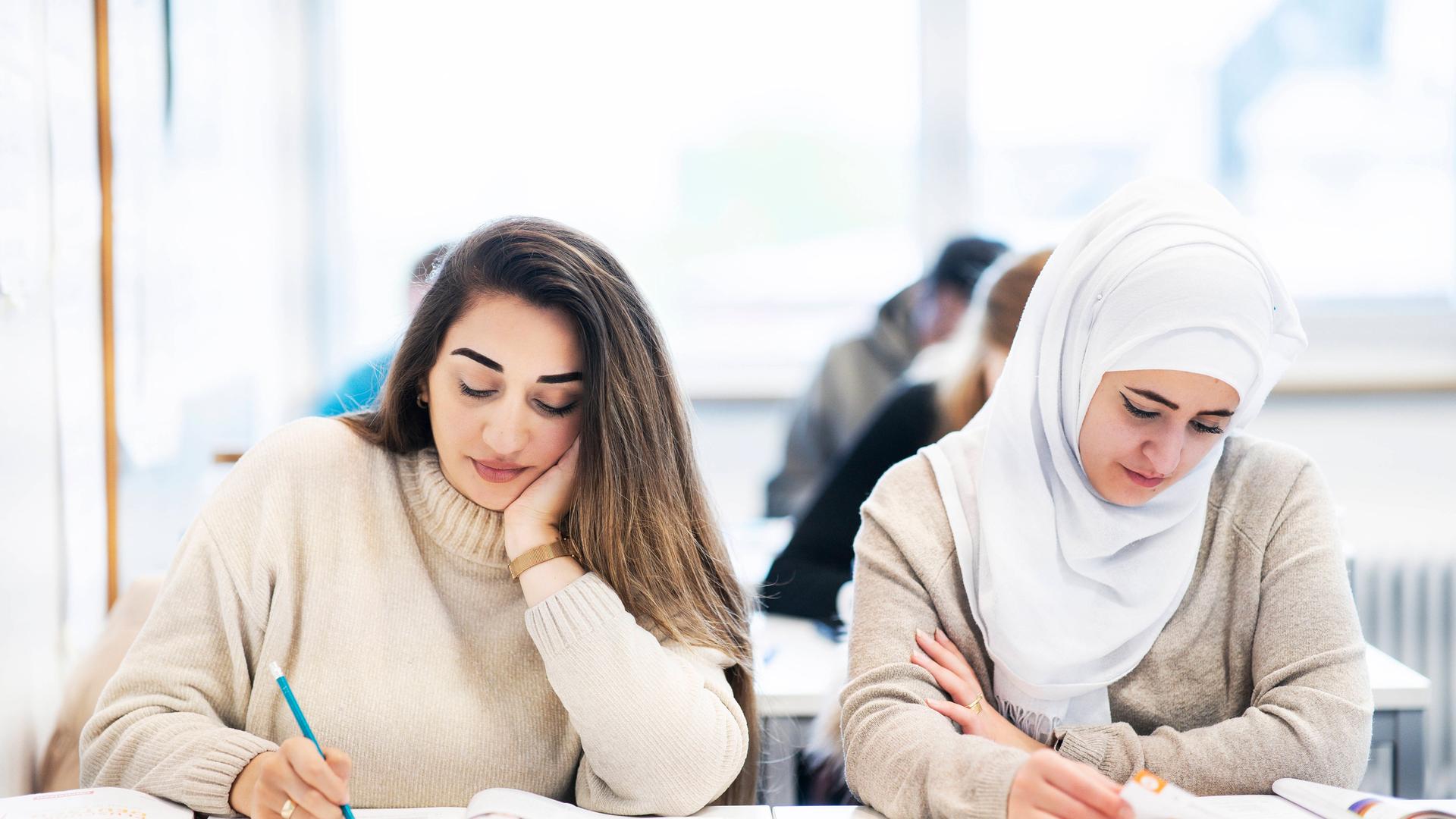 Zwei junge Frauen, eine von ihnen mit Kopftuch in einem Klassenzimmer schreiben etwas in ein Heft. Zwei junge Frauen, eine von ihnen mit Kopftuch in einem Klassenzimmer schreiben etwas in ein Heft.