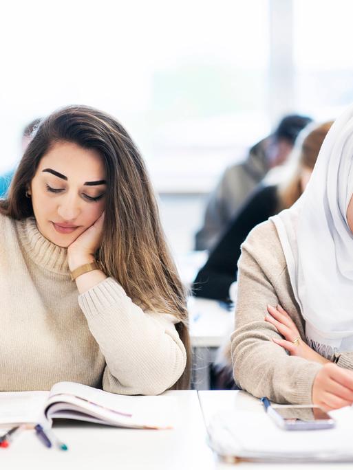 Zwei junge Frauen, eine von ihnen mit Kopftuch in einem Klassenzimmer schreiben etwas in ein Heft.