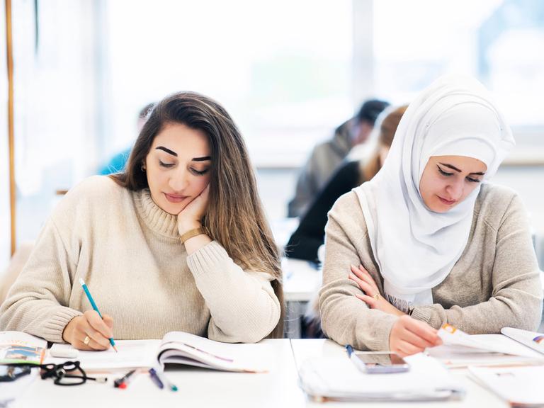 Zwei junge Frauen, eine von ihnen mit Kopftuch in einem Klassenzimmer schreiben etwas in ein Heft.