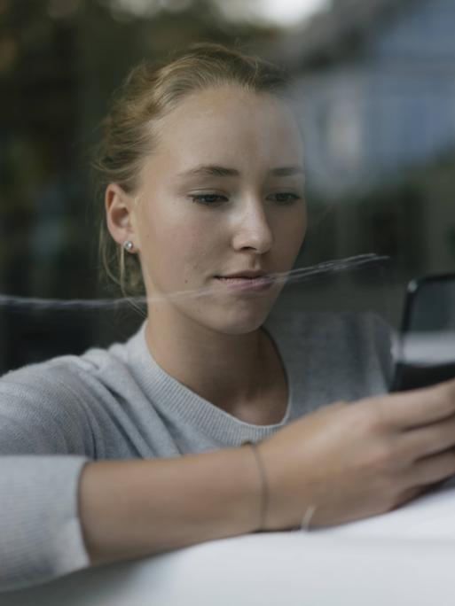Eine junge Frau sitzt auf einem Sofa, hinter einer Fensterscheibe. Sie hält mit beiden Händen ein Smartphone und blickt darauf.