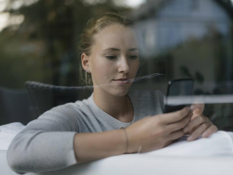 Eine junge Frau sitzt auf einem Sofa, hinter einer Fensterscheibe. Sie hält mit beiden Händen ein Smartphone und blickt darauf.