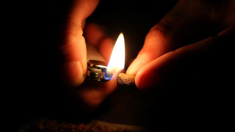 Zwei Hände im Dunkeln: Die eine Hand hält ein Stück Marihuana, die andere ein Feuerzeug mit Flamme. Zwei Hände im Dunkeln: Die eine Hand hält ein Stück Marihuana, die andere ein Feuerzeug mit Flamme.