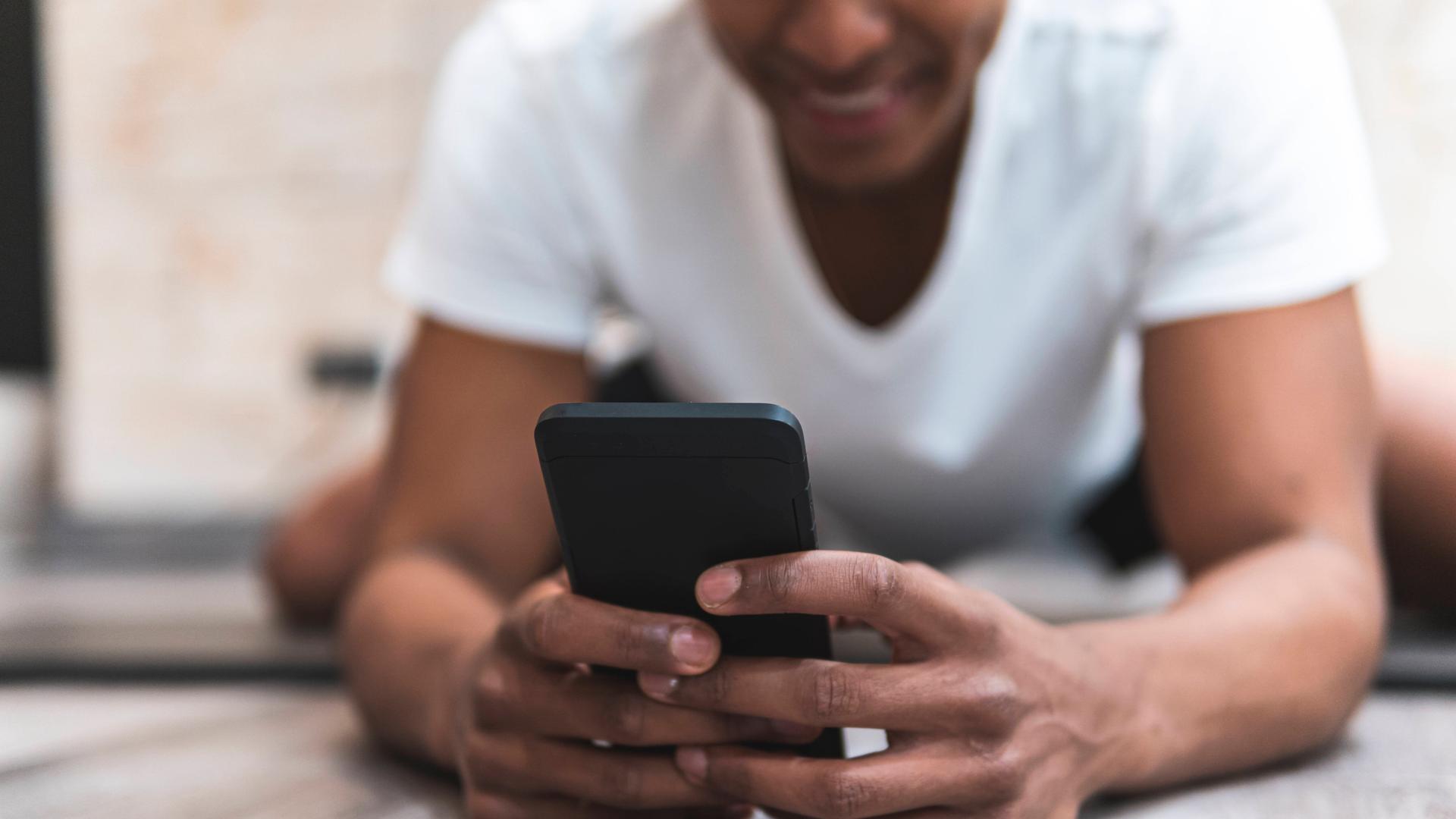 Foto eines jungen Mannes, der auf dem Fußboden liegend auf ein Smartphone guckt