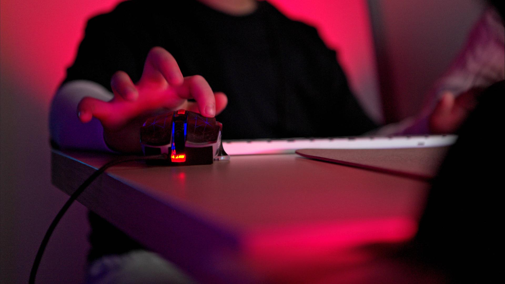 Nahaufnahme einer Person, die eine Gaming-Maus mit RGB-Beleuchtung neben einer Tastatur in einem dunklen Raum mit neonrotem Licht verwendet. Die Person ist nicht zu sehen, lediglich Oberkörper und die Hand auf der Gaming-Maus.