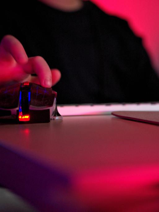 Nahaufnahme einer Person, die eine Gaming-Maus mit RGB-Beleuchtung neben einer Tastatur in einem dunklen Raum mit neonrotem Licht verwendet. Die Person ist nicht zu sehen, lediglich Oberkörper und die Hand auf der Gaming-Maus.