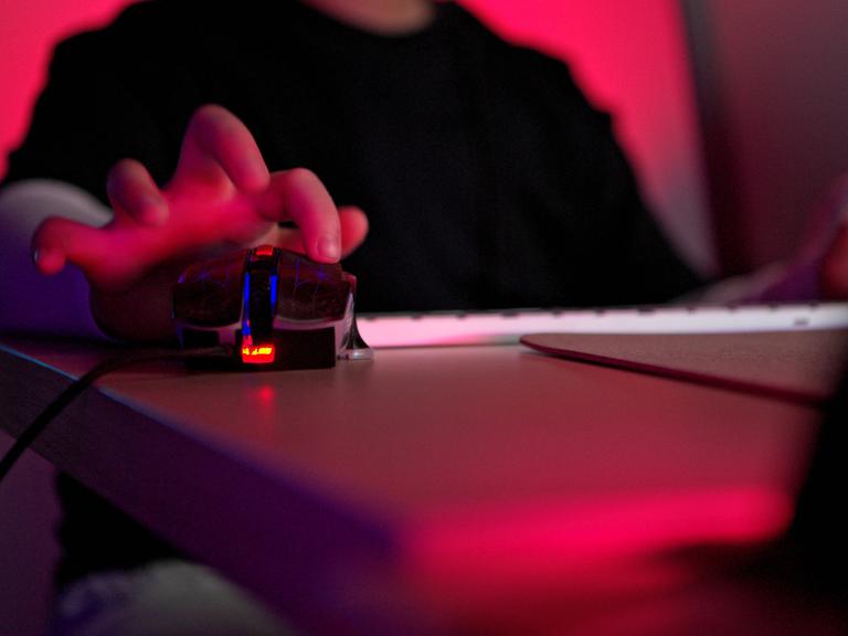 Nahaufnahme einer Person, die eine Gaming-Maus mit RGB-Beleuchtung neben einer Tastatur in einem dunklen Raum mit neonrotem Licht verwendet. Die Person ist nicht zu sehen, lediglich Oberkörper und die Hand auf der Gaming-Maus.