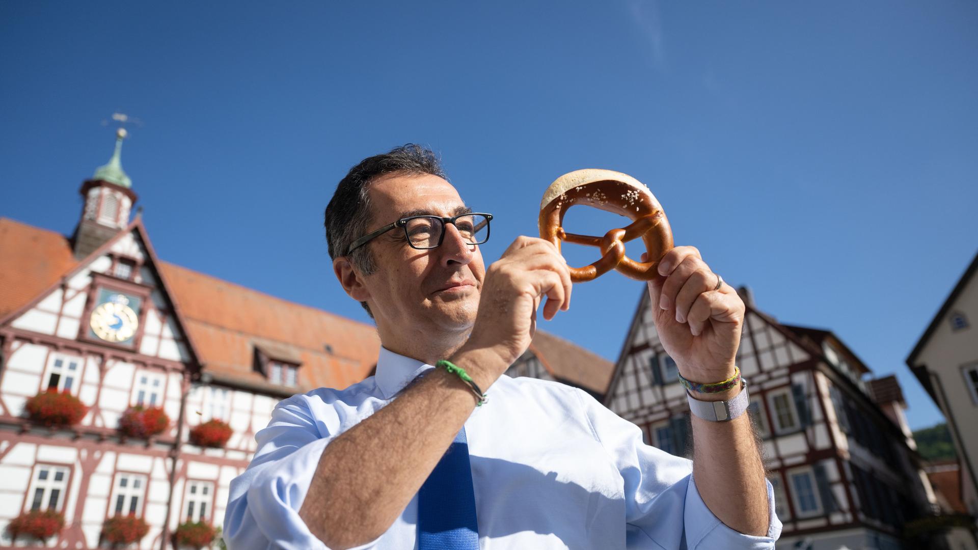 Bundesernährungsminister Cem Özdemir (Bündnis 90/Die Grünen) zeigt auf dem Marktplatz seines Geburtsorts nach dem Besuch einer Bäckerei eine Brezel.