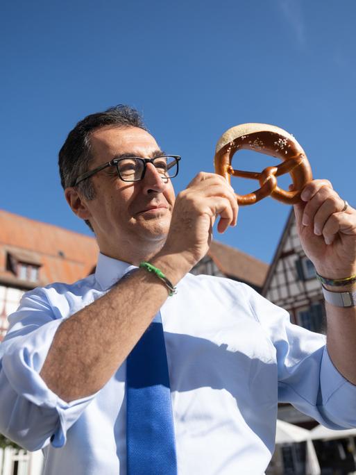 Bundesernährungsminister Cem Özdemir (Bündnis 90/Die Grünen) zeigt auf dem Marktplatz seines Geburtsorts nach dem Besuch einer Bäckerei eine Brezel.