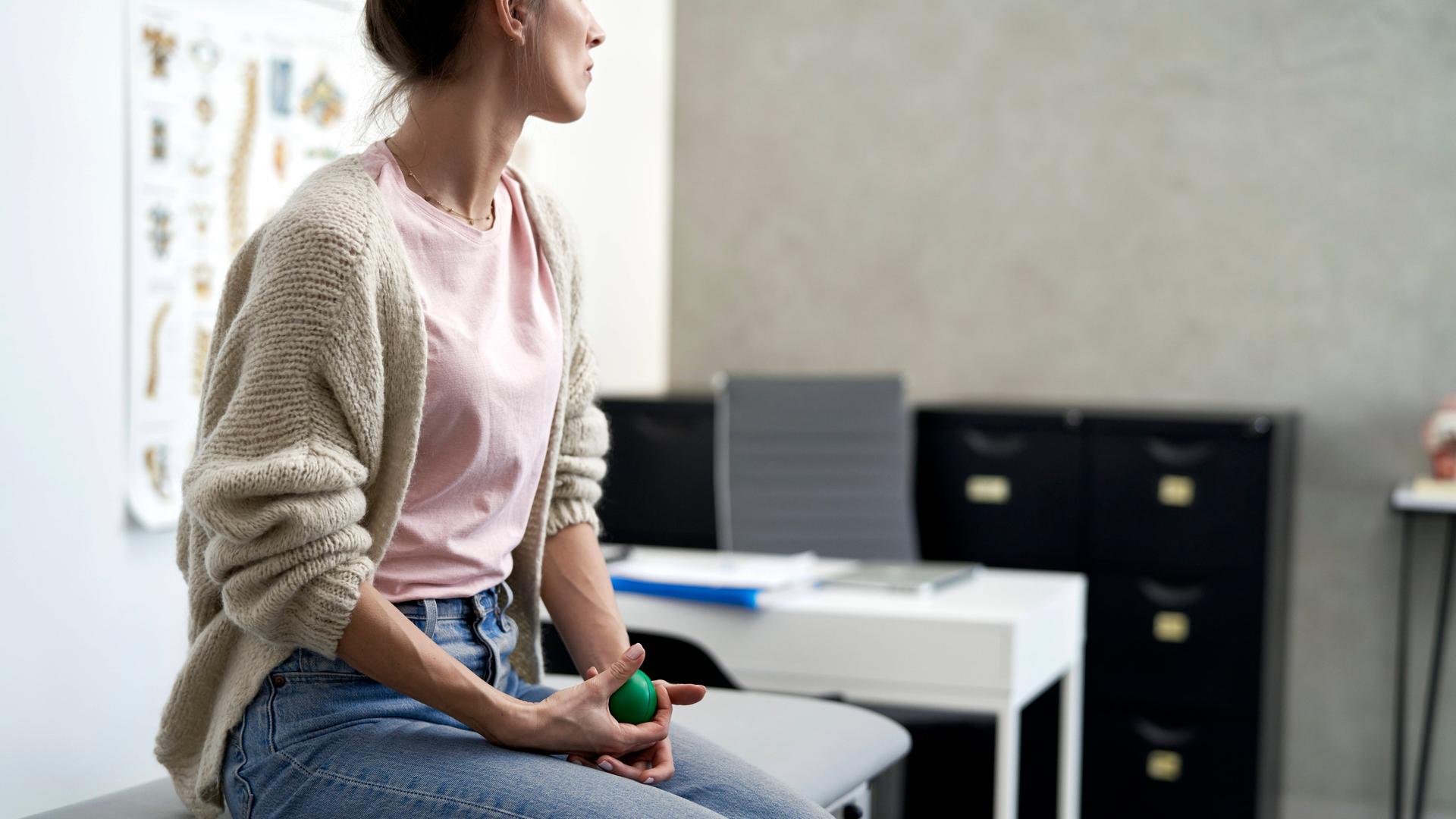 Eine Frau sitzt in einem Behandlungszimmer beim Arzt auf einer Liege. Sie wartet dort und hält dabei einen kleinen grünen Gummiball in den Händen.