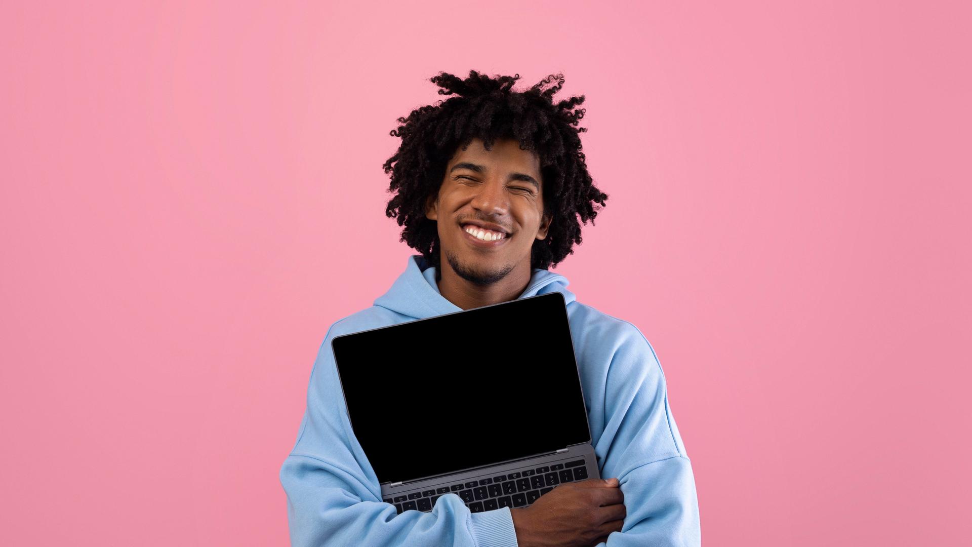 Foto eines jungen Mannes in blauem Pullover vor einer pinken Wand, der ein aufgeklapptes Laptop umarmt