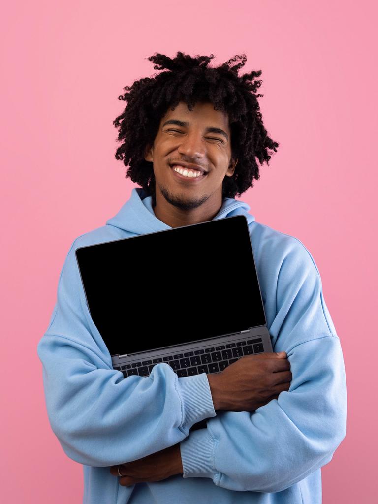 Foto eines jungen Mannes in blauem Pullover vor einer pinken Wand, der ein aufgeklapptes Laptop umarmt