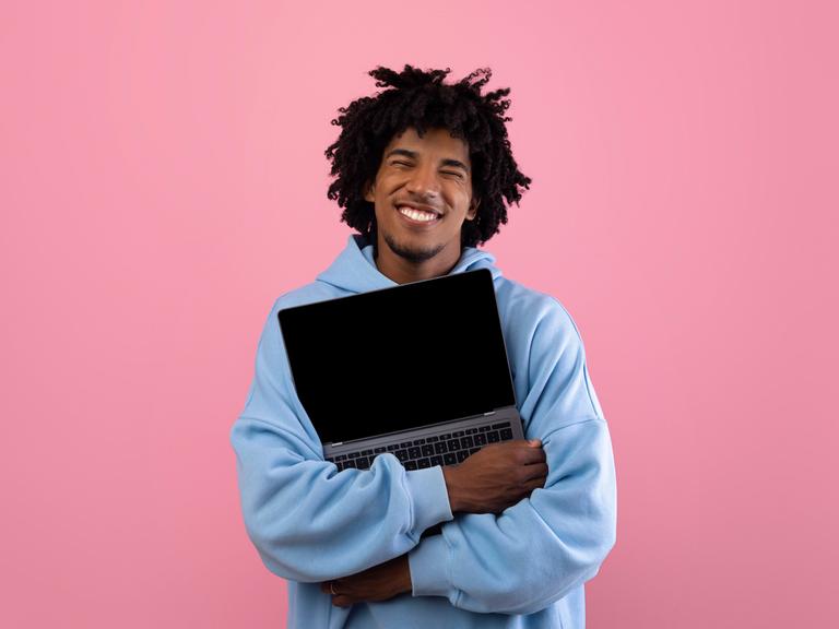 Foto eines jungen Mannes in blauem Pullover vor einer pinken Wand, der ein aufgeklapptes Laptop umarmt