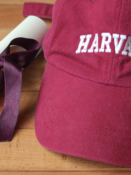 Eine bordeauxfarbene Baseballkappe mit Harvard-Logo und einem eingerollten Dokument, das von einer violetten Schleife zusammengehalten wird. Eine bordeauxfarbene Baseballkappe mit Harvard-Logo und einem eingerollten Dokument, das von einer violetten Schleife zusammengehalten wird.