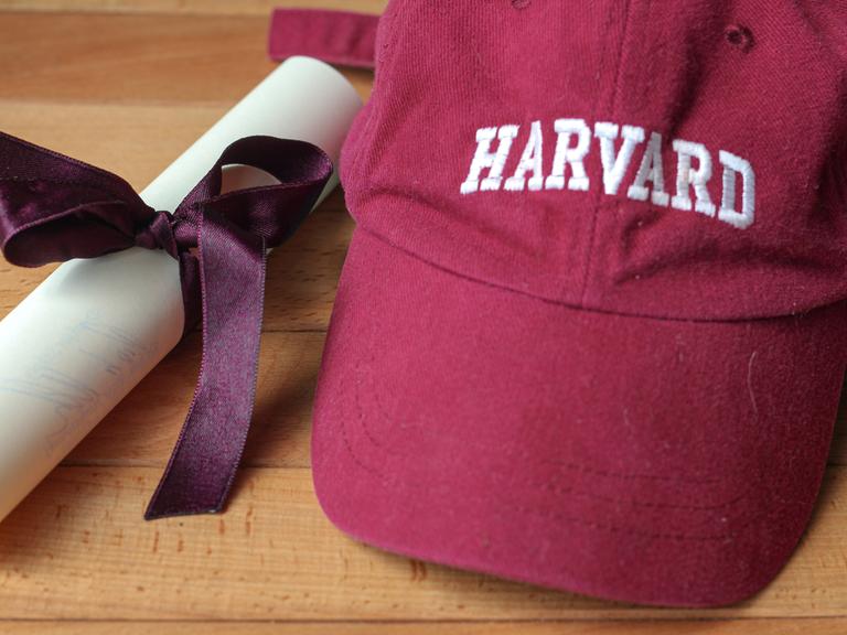 Eine bordeauxfarbene Baseballkappe mit Harvard-Logo und einem eingerollten Dokument, das von einer violetten Schleife zusammengehalten wird. Eine bordeauxfarbene Baseballkappe mit Harvard-Logo und einem eingerollten Dokument, das von einer violetten Schleife zusammengehalten wird.