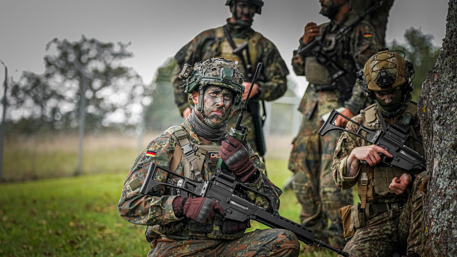 Vier Bundeswehrsoldaten tragen bei einer Übung Unform, Waffen, haben ihre Gesichter mit Tarnfarbe geschminkt und Stellung an einem Baum auf einer Wiese bezogen.