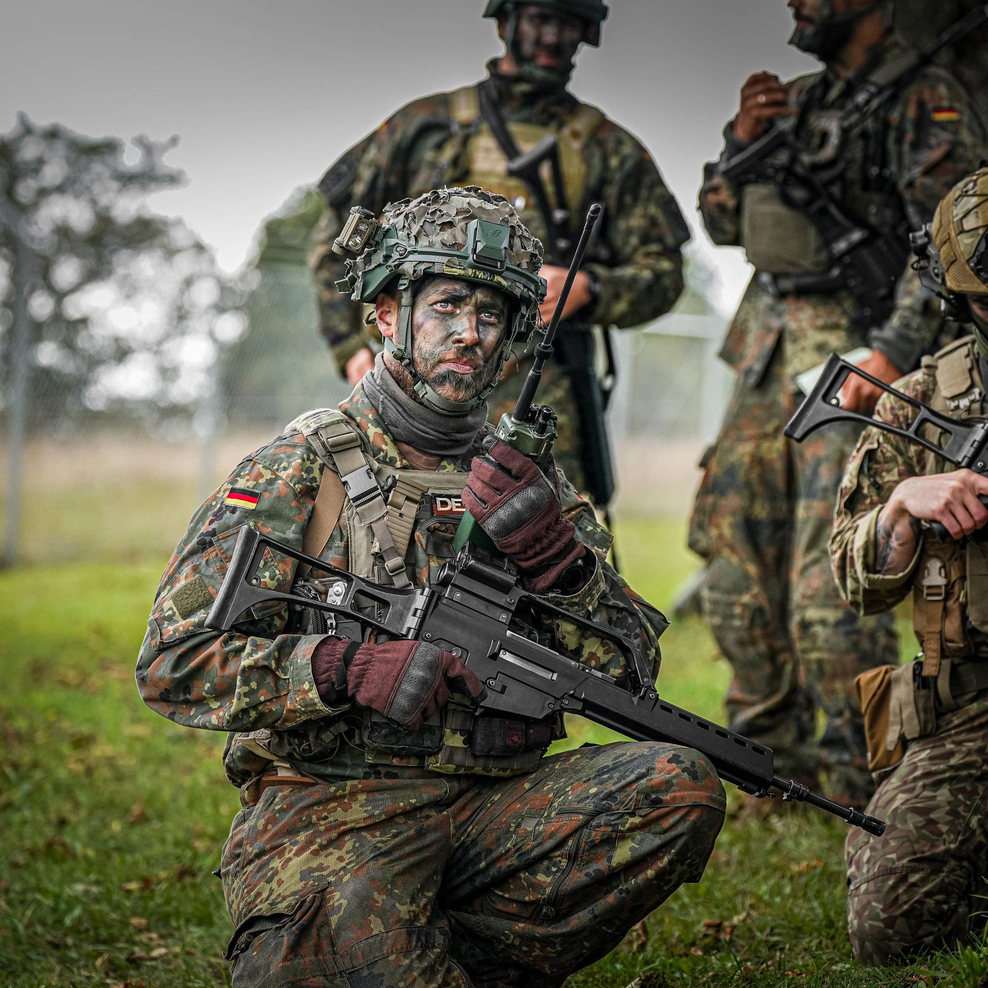 Vier Bundeswehrsoldaten tragen bei einer Übung Unform, Waffen, haben ihre Gesichter mit Tarnfarbe geschminkt und Stellung an einem Baum auf einer Wiese bezogen.