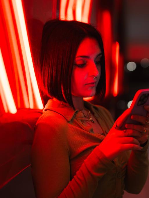 Eine Frau lehnt an die Glasfassade eines Gebäudes und schaut auf ihr Handy. Hinter dem Glas sind rote Neonröhren eingebaut, die die Frau und das Gebäude in rotes Licht umhüllen.