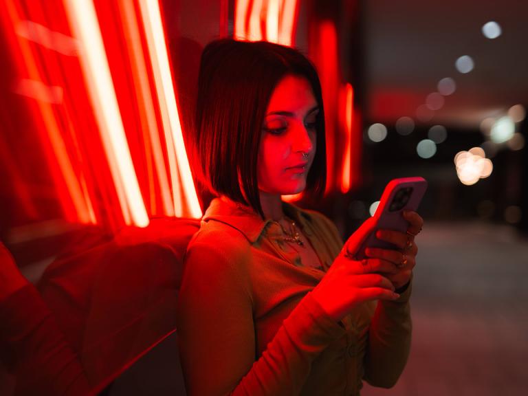 Eine Frau lehnt an die Glasfassade eines Gebäudes und schaut auf ihr Handy. Hinter dem Glas sind rote Neonröhren eingebaut, die die Frau und das Gebäude in rotes Licht umhüllen.
