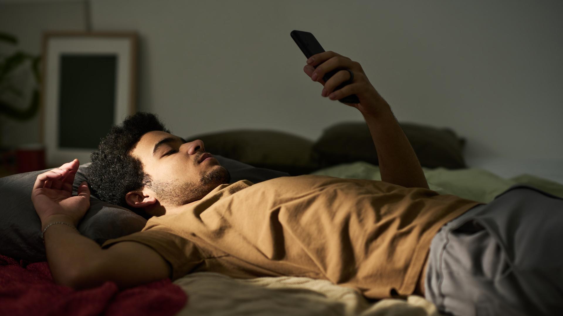Ein Mann liegt auf dem Rücken im Bett. Sein linker Arm ist aufgestützt und er hält ein Smartphone in die Höhe, auf das er blickt. Es wirkt ruhig und etwas traurig. Ein Mann liegt auf dem Rücken im Bett. Sein linker Arm ist aufgestützt und er hält ein Smartphone in die Höhe, auf das er blickt. Es wirkt ruhig und etwas traurig.