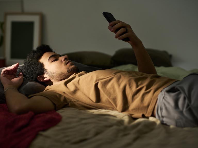 Ein Mann liegt auf dem Rücken im Bett. Sein linker Arm ist aufgestützt und er hält ein Smartphone in die Höhe, auf das er blickt. Es wirkt ruhig und etwas traurig.