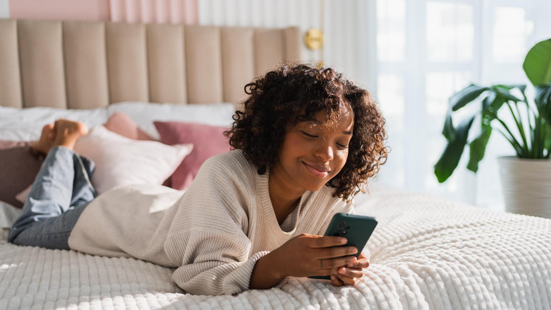 Foto einer jungen Frau, die lächelnd auf dem Bett liegt und auf ihrem Smartphone tippt
