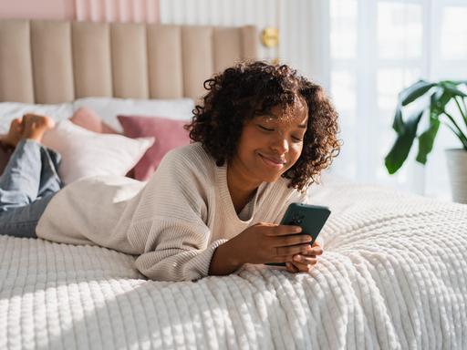 Foto einer jungen Frau, die lächelnd auf dem Bett liegt und auf ihrem Smartphone tippt