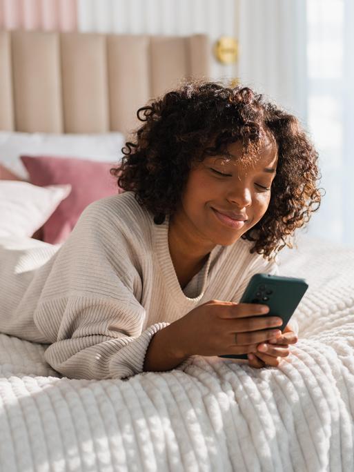 Foto einer jungen Frau, die lächelnd auf dem Bett liegt und auf ihrem Smartphone tippt