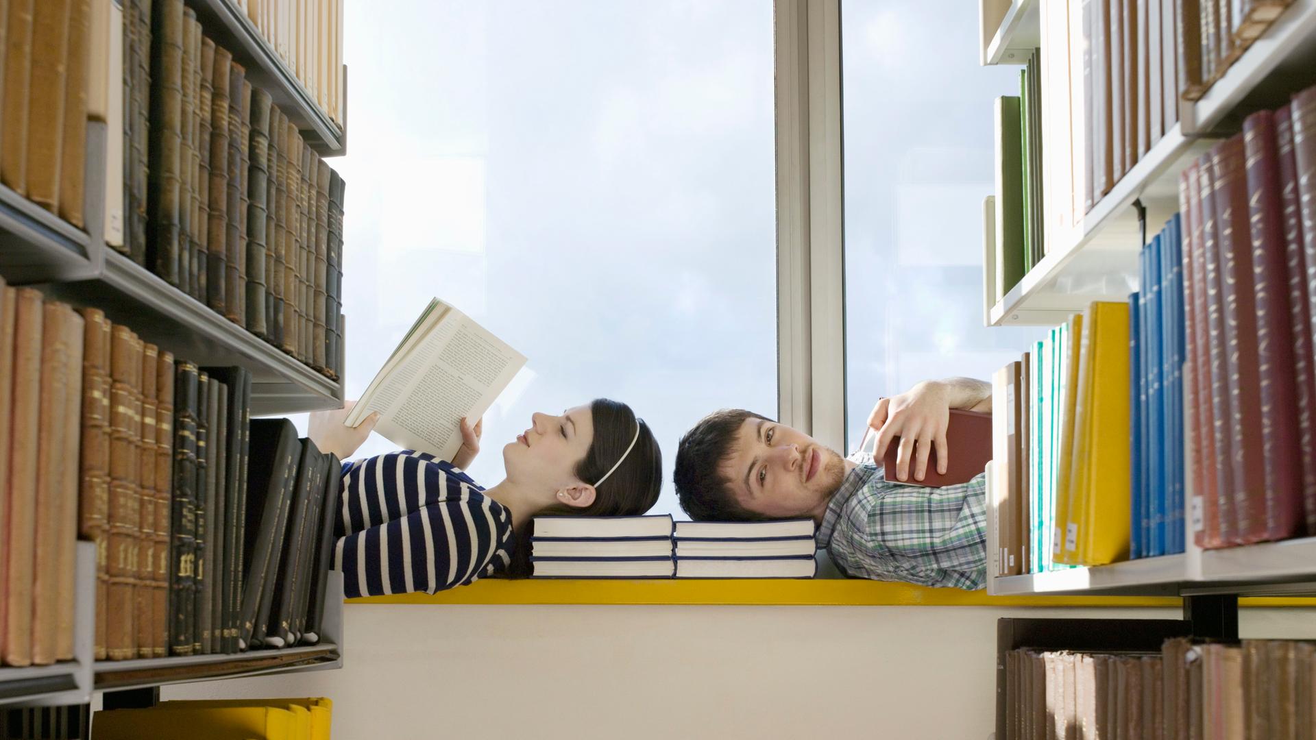 Zwei Personen liegen mit den Köpfen zueinander auf einer Fensterbank in einer Bibliothek