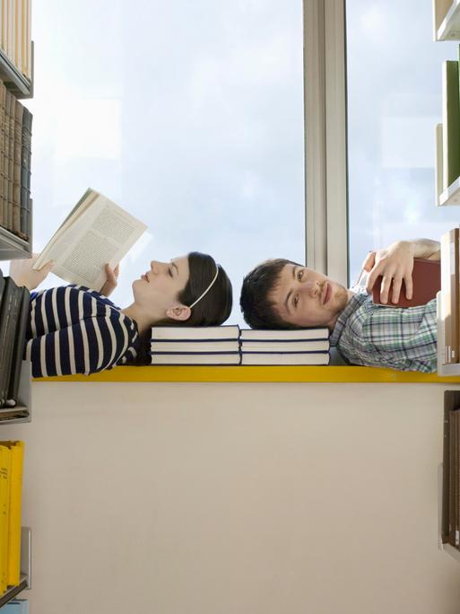 Zwei Personen liegen mit den Köpfen zueinander auf einer Fensterbank in einer Bibliothek