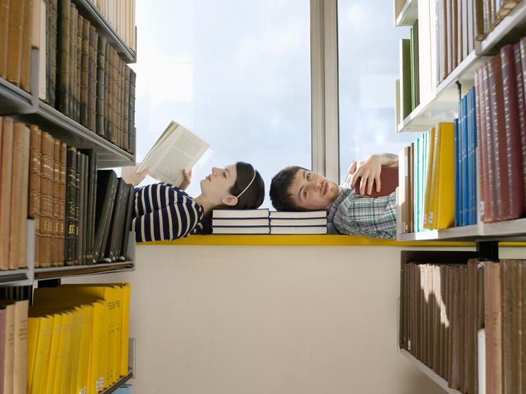 Zwei Personen liegen mit den Köpfen zueinander auf einer Fensterbank in einer Bibliothek