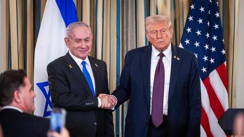 Netanjahu und Trump reichen sich die Hand. Im Hintergrund die Flaggen Israels und der USA. Reporter fotografieren die Szene.