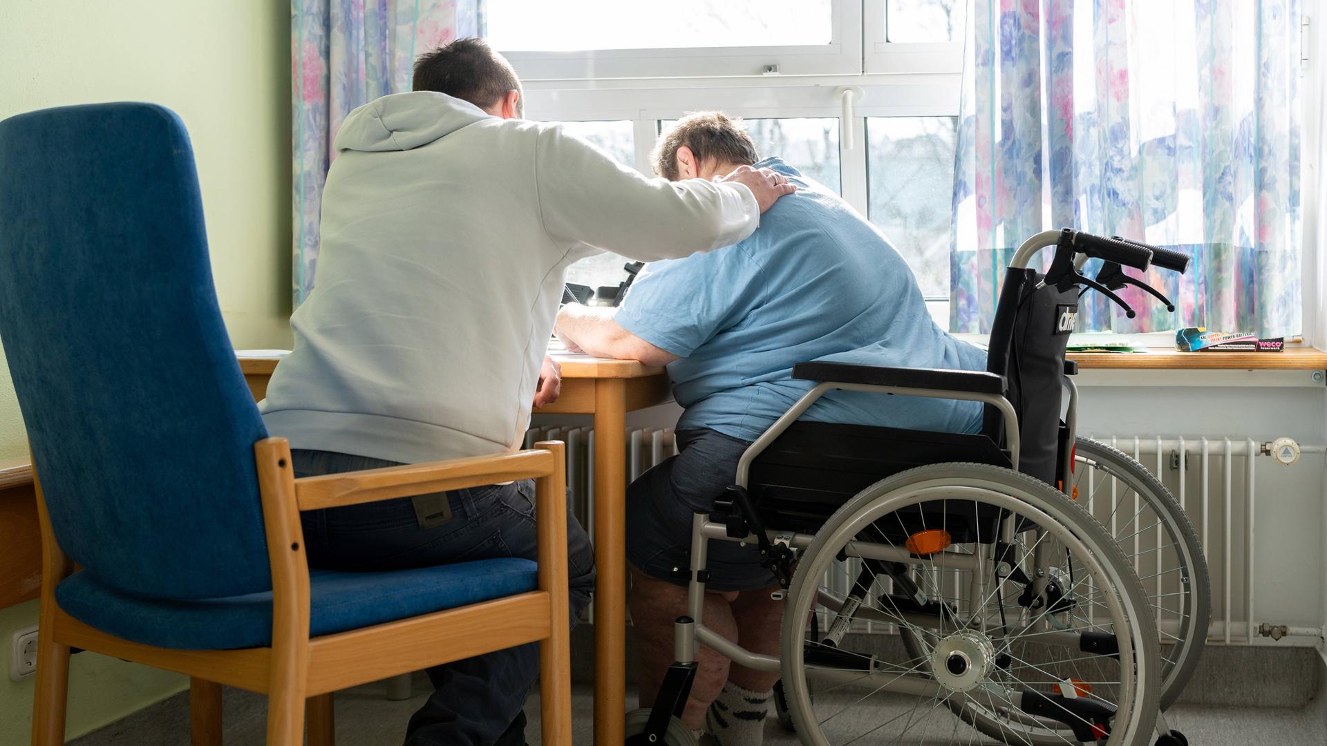 Ein Mann betreut eine Frau im Rollstuhl und legt ihr den Arm auf die Schulter.