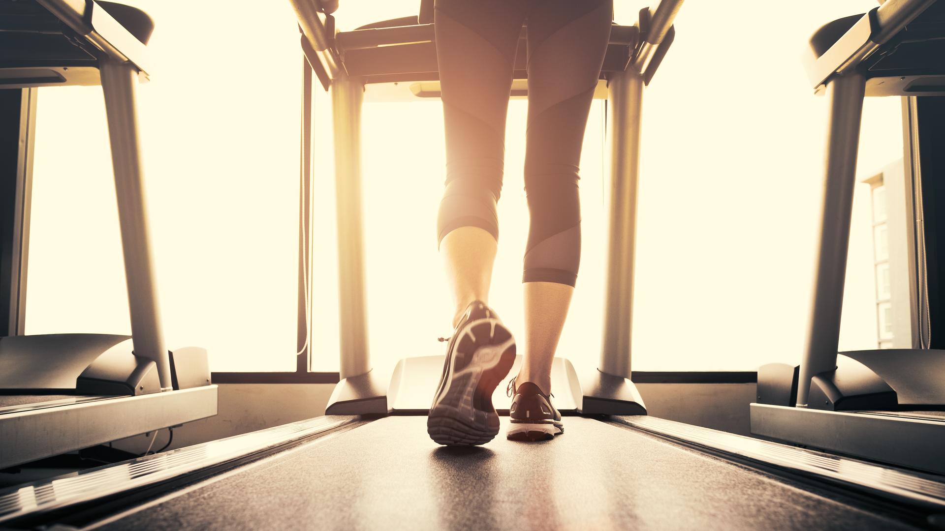 Eine Frau steht auf einem Laufband und läuft; sie trägt Turnschuhe und eine Leggings. Sie ist von hinten zu sehen. Durch das Fenster vor ihr fällt Sonnenlicht. Eine Frau steht auf einem Laufband und läuft; sie trägt Turnschuhe und eine Leggings. Sie ist von hinten zu sehen. Durch das Fenster vor ihr fällt Sonnenlicht.