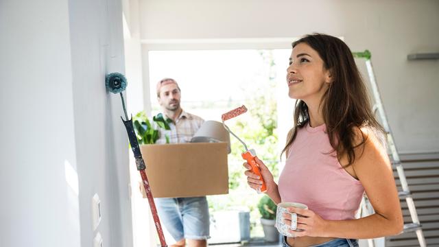 Eine Frau hat eine Farbrolle in der Hand und lächelt, im Hintergrund trägt ein Mann einen Umzugskarton