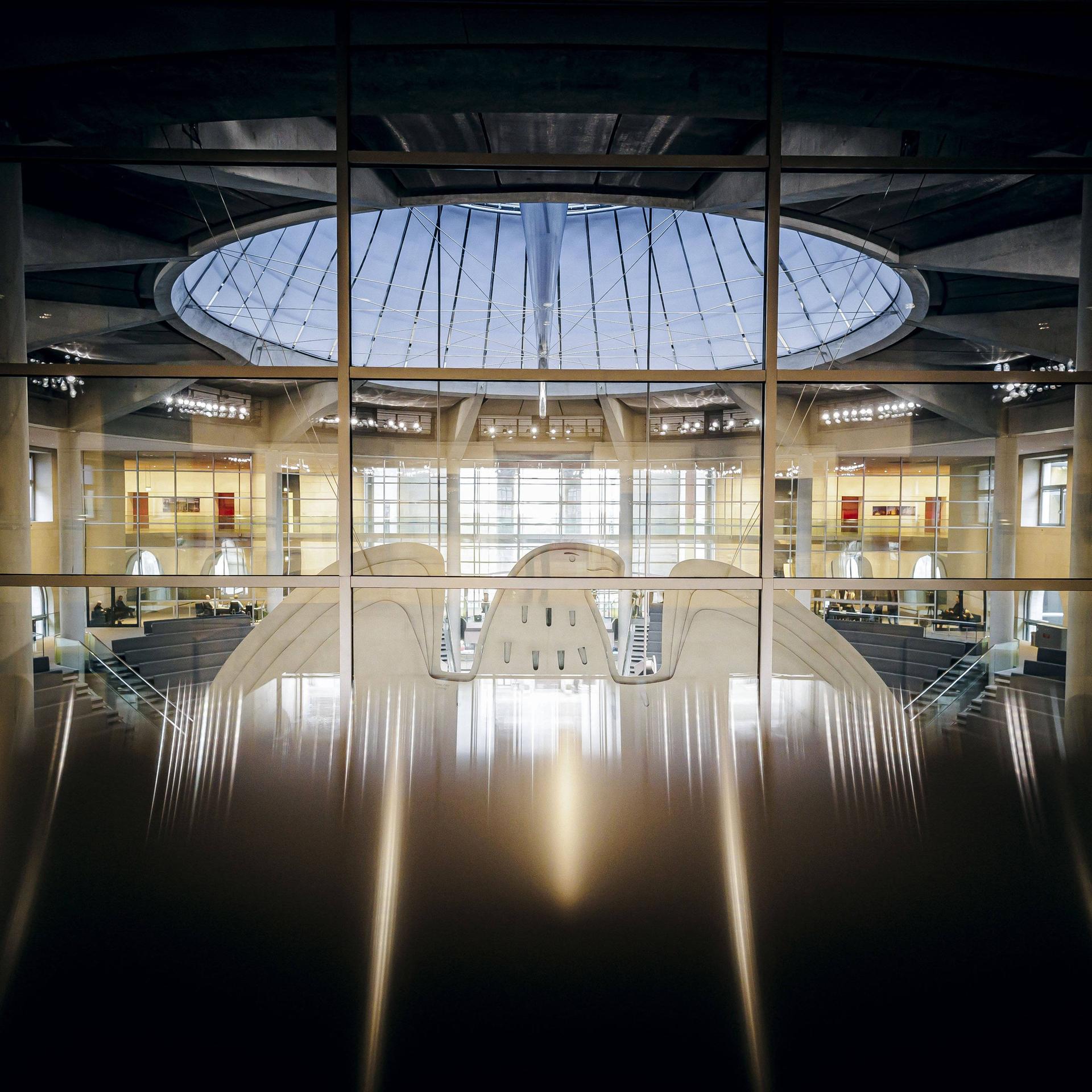 Blick durch eine Glasfront von hinten auf den Bundesadler im Plenum des Bundestages.