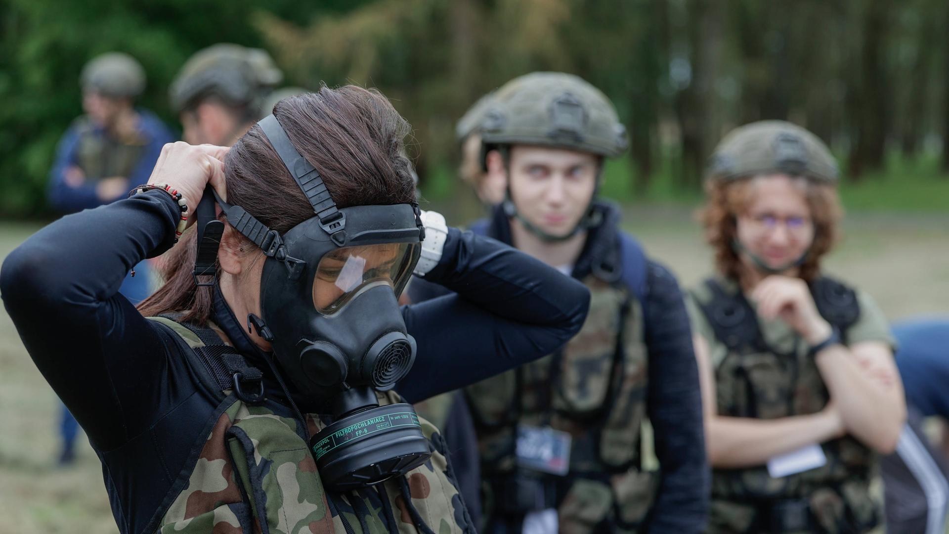 Eine junge Frau probiert eine Gasmaske an. Im Hintergrund stehen junge Menschen in Flecktarn, die sie ansehen Eine junge Frau probiert eine Gasmaske an. Im Hintergrund stehen junge Menschen in Flecktarn, die sie ansehen