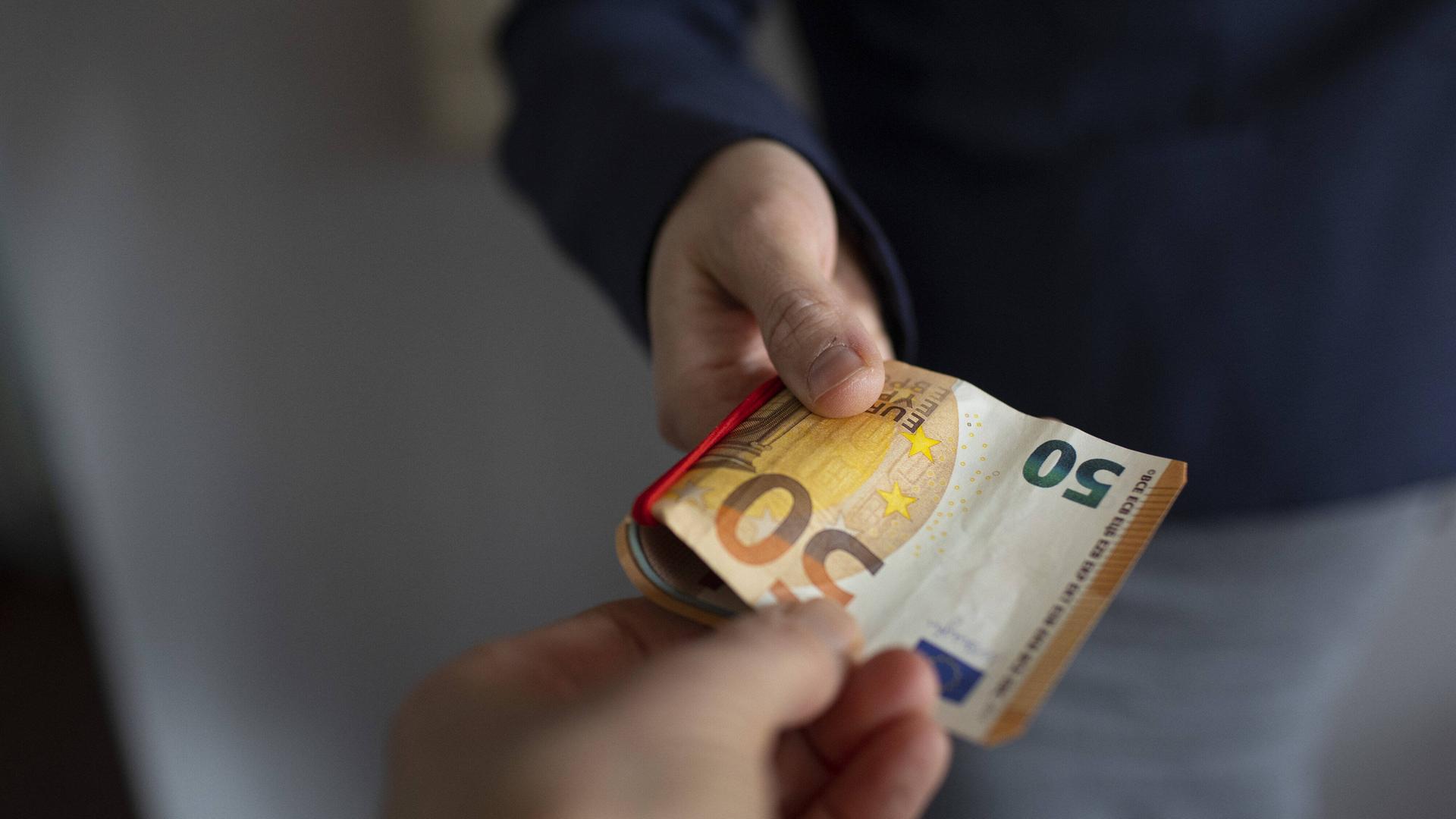Eine Person hält einen 50-Euro-Schein in der Hand und streckt ihn einer anderen Person hin.