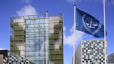 Mehrere große Gebäude mit Glasfront, dazwischen weht eine blaue Flagge mit dem Logo des Internationalen Strafgerichtshofs.
