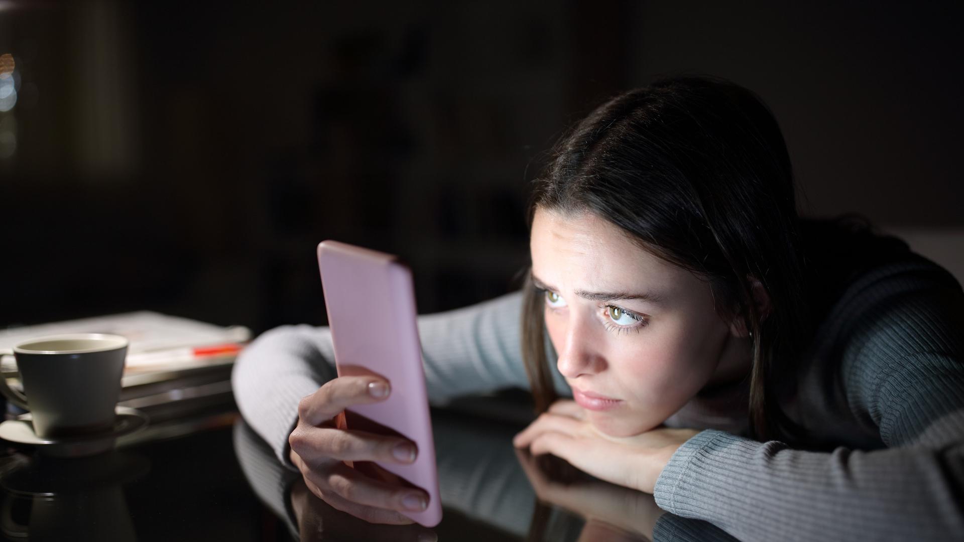 Eine junge Frau schaut auf ein Smartphone; sie stützt sich auf einem Tisch ab und hat den Kopf auf eine Hand gelegt. Eine junge Frau schaut auf ein Smartphone; sie stützt sich auf einem Tisch ab und hat den Kopf auf eine Hand gelegt.