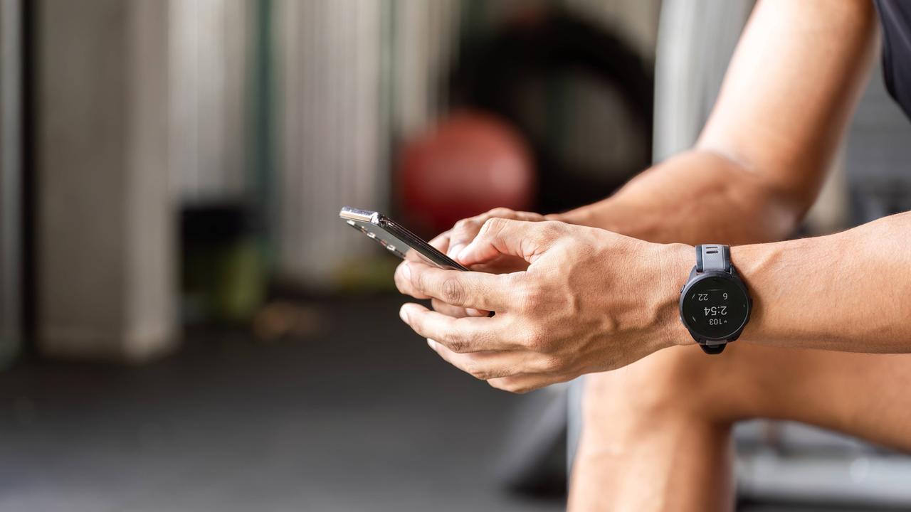 Ein Sportler nutzt eine Fitness-App auf seinem Smartphone während einer Pause im Fitnessstudio. Er sitzt dort, konzentriert auf sein Smartphone und verfolgt mithilfe einer Fitness-App seine Fortschritte und Trainingseinheiten.