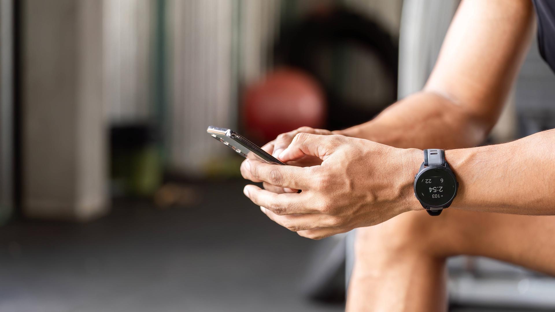 Ein Sportler nutzt eine Fitness-App auf seinem Smartphone während einer Pause im Fitnessstudio. Er sitzt dort, konzentriert auf sein Smartphone und verfolgt mithilfe einer Fitness-App seine Fortschritte und Trainingseinheiten.