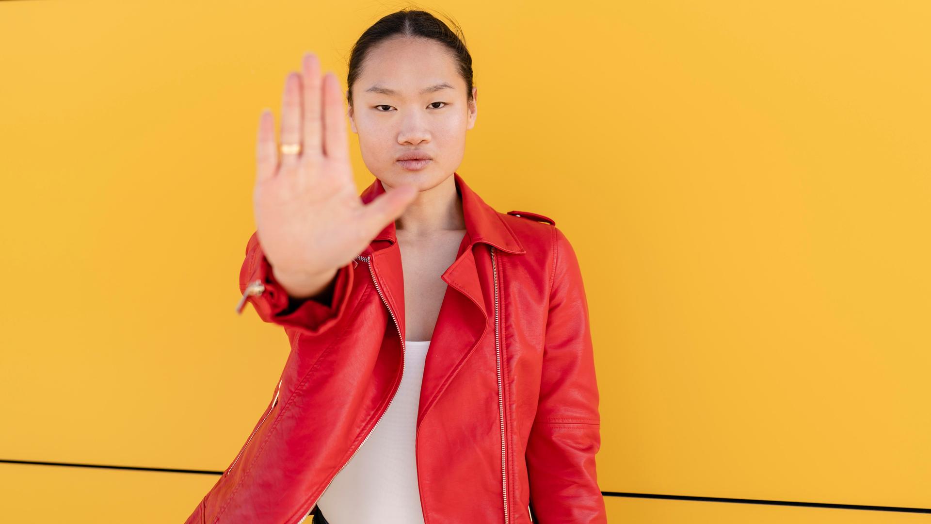 Eine Frau steht vor einer gelben Wand. Sie trägt eine rote Lederjacke, schaut in die Kamera und hält ihre flache Hand hoch zu einer Stopp-Geste.