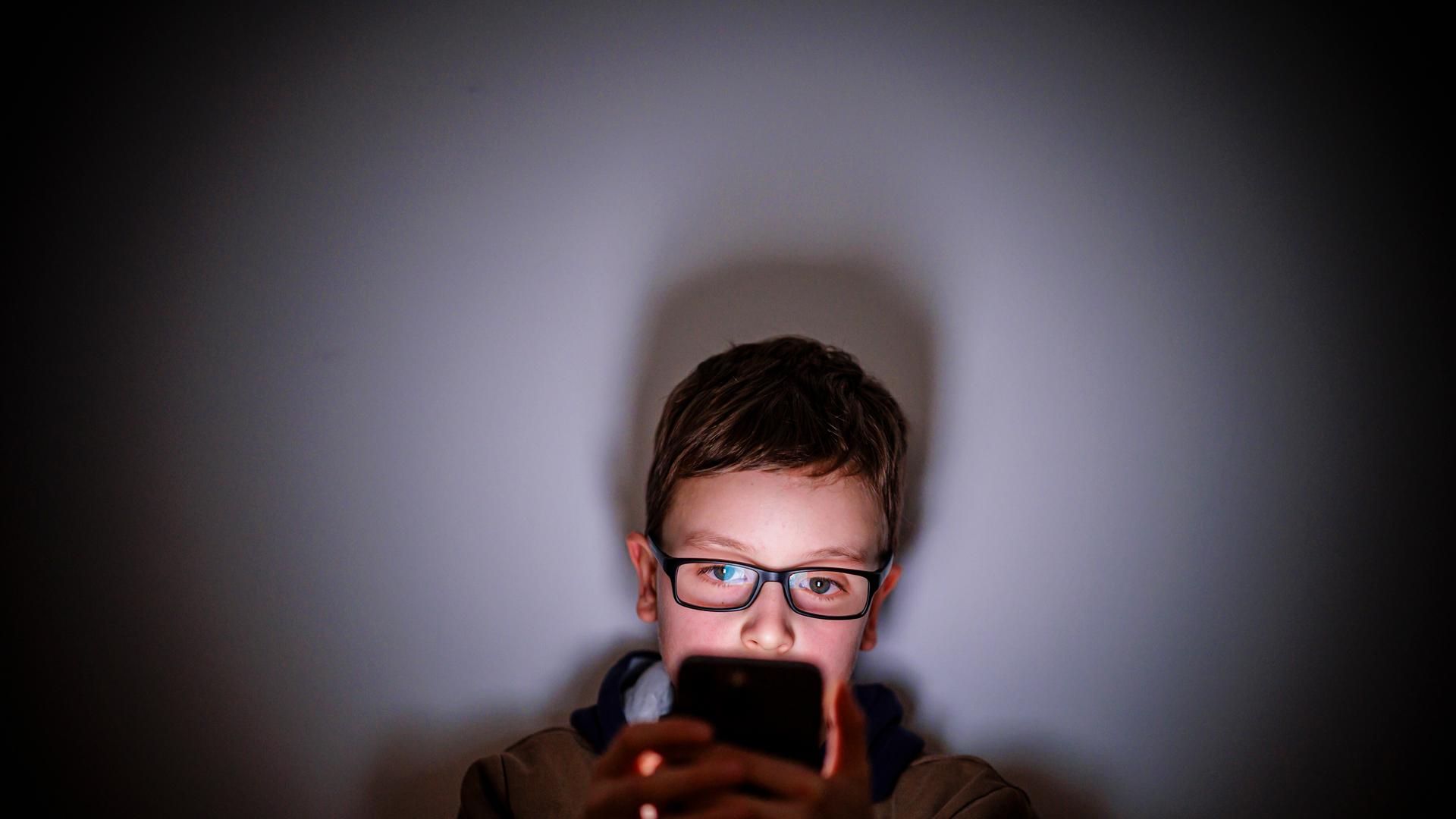 Ein Junge sitzt in einem dunklen Raum und wird von einem Smartphone beleuchtet. Symbolfoto. Gestellte Aufnahme zum Thema Kinder nutzen Social Media.