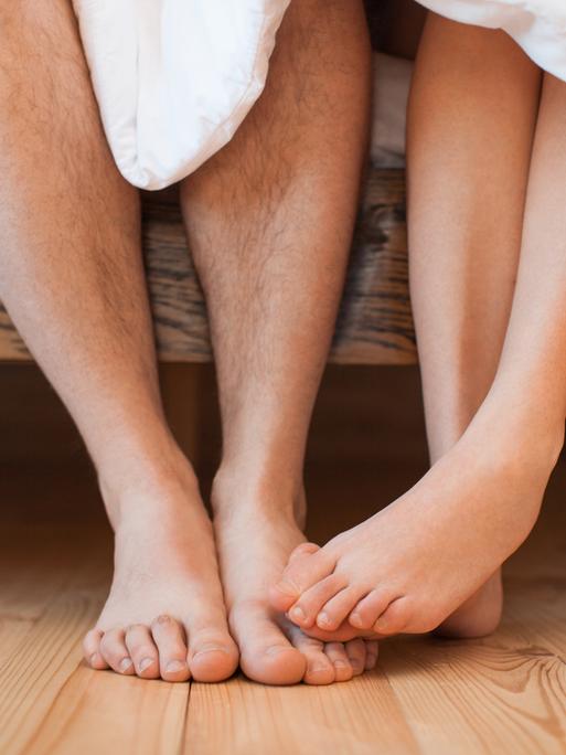 Ein junges Paar sitzt bafüßig und unbekleidet mit einer Decke bedecktauf einem Bett Ein junges Paar sitzt bafüßig und unbekleidet mit einer Decke bedecktauf einem Bett