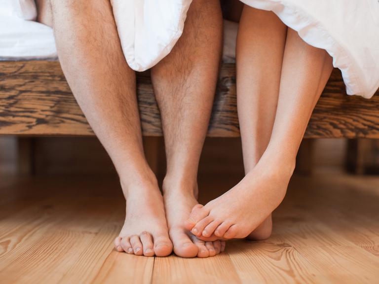 Ein junges Paar sitzt bafüßig und unbekleidet mit einer Decke bedecktauf einem Bett Ein junges Paar sitzt bafüßig und unbekleidet mit einer Decke bedecktauf einem Bett