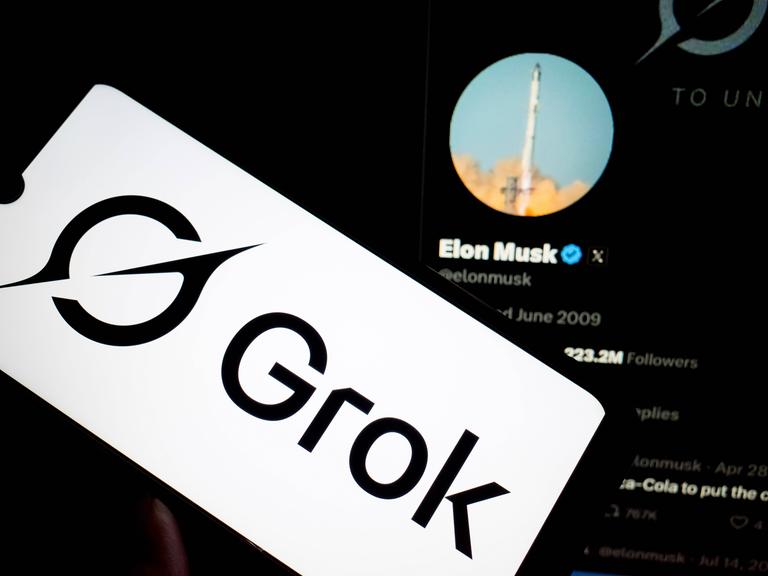 Das Logo der KI Grok von X (ehemals Twitter) auf einem Smartphone-Display. Im Hintergrund das X-Profil des X-Eigentümers Elon Musk auf einem größeren Display.
