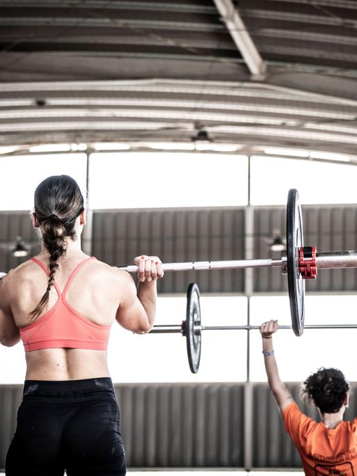 Eine Frau hebt mit ihren Armen Gewichte an einer Langhantel nach oben. Eine Frau hebt mit ihren Armen Gewichte an einer Langhantel nach oben.