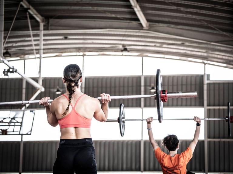 Eine Frau hebt mit ihren Armen Gewichte an einer Langhantel nach oben. Eine Frau hebt mit ihren Armen Gewichte an einer Langhantel nach oben.