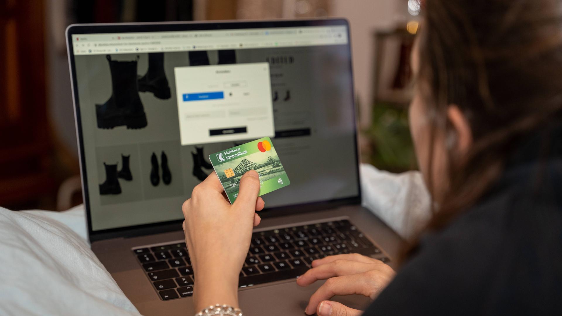 Eine Frau sitzt vor einem Laptop und hat eine Shoppingseite geöffnet. in der linken Hand hält sie ihre Kreditkarte, während sie mit der rechten tippt.